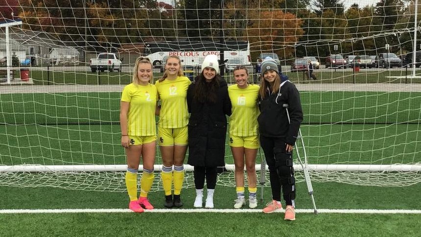 5 women's soccer players standing in front of the soccer goal smiling for a picture